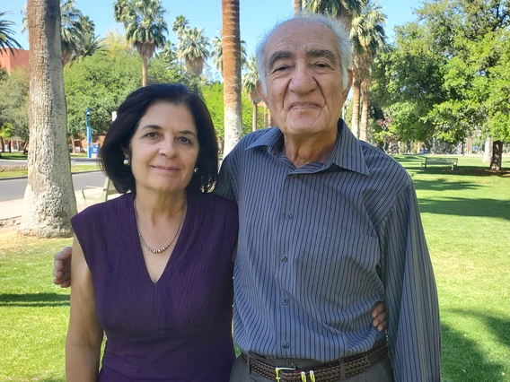 two people, standing and smiling, palm trees and grass in background