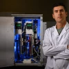 A man in a white lab coat stand in front of a blue-lit machine.
